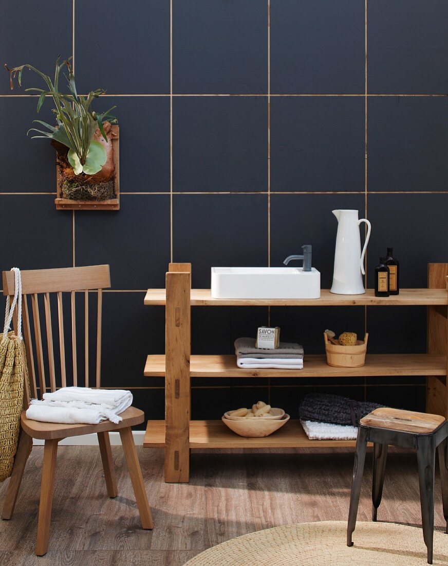 Bathroom utensils on simple, plain wood shelves against dark blue tiles wall with wooden chair and stool in foreground