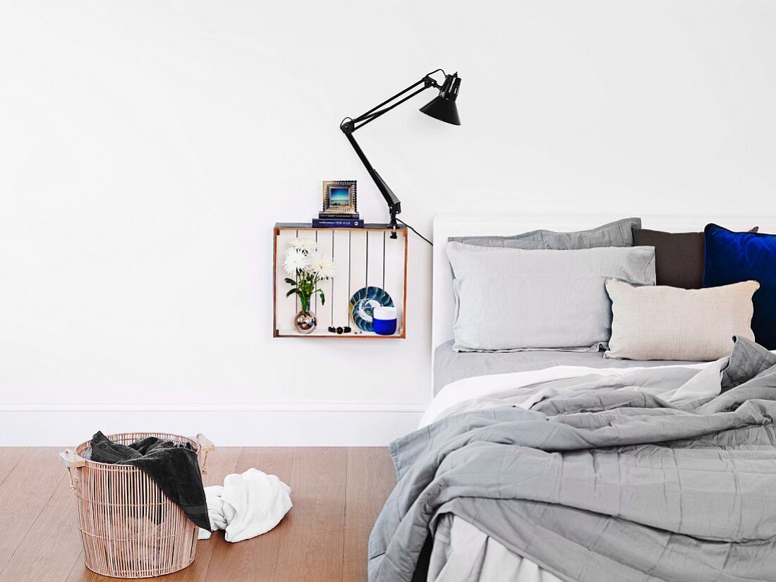 White wooden crate mounted on wall and painted white inside with ornaments and clip lamp used as bedside cabinet next to unmade bed and laundry basket