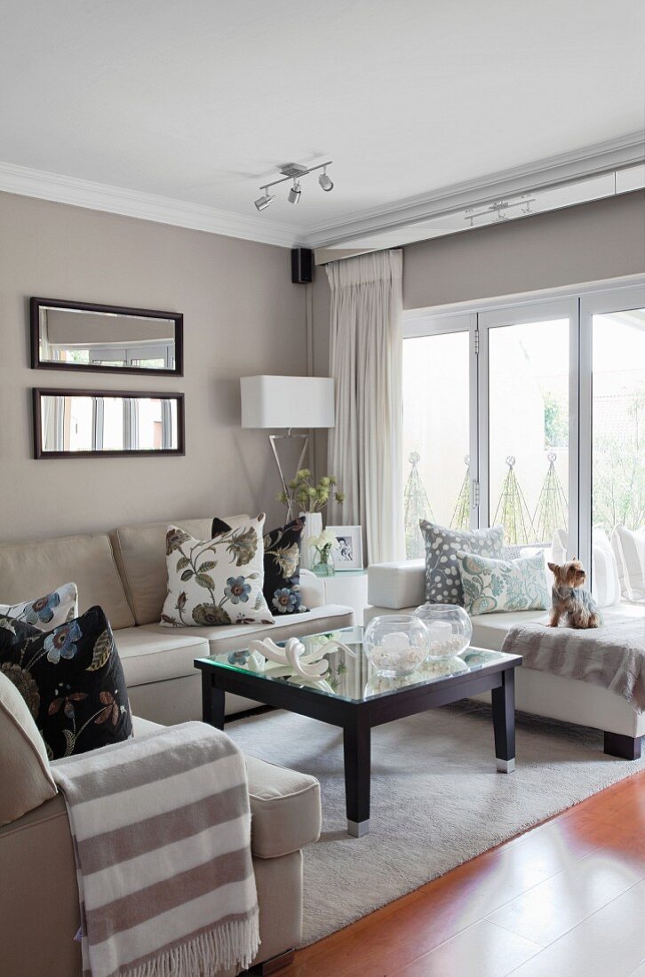 Framed, landscape-format mirrors and mirrored curtain rails in seating area with patterned scatter cushions and blanket with wide stripes on beige sofas