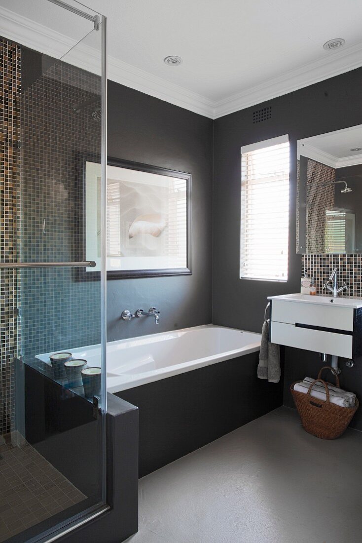 Dunkle, unifarbene Wände im Badewannenbereich ergänzt mit Mosaikfliesen hinter dem Waschtisch und der spiegelnden Glastür der Duschkabine