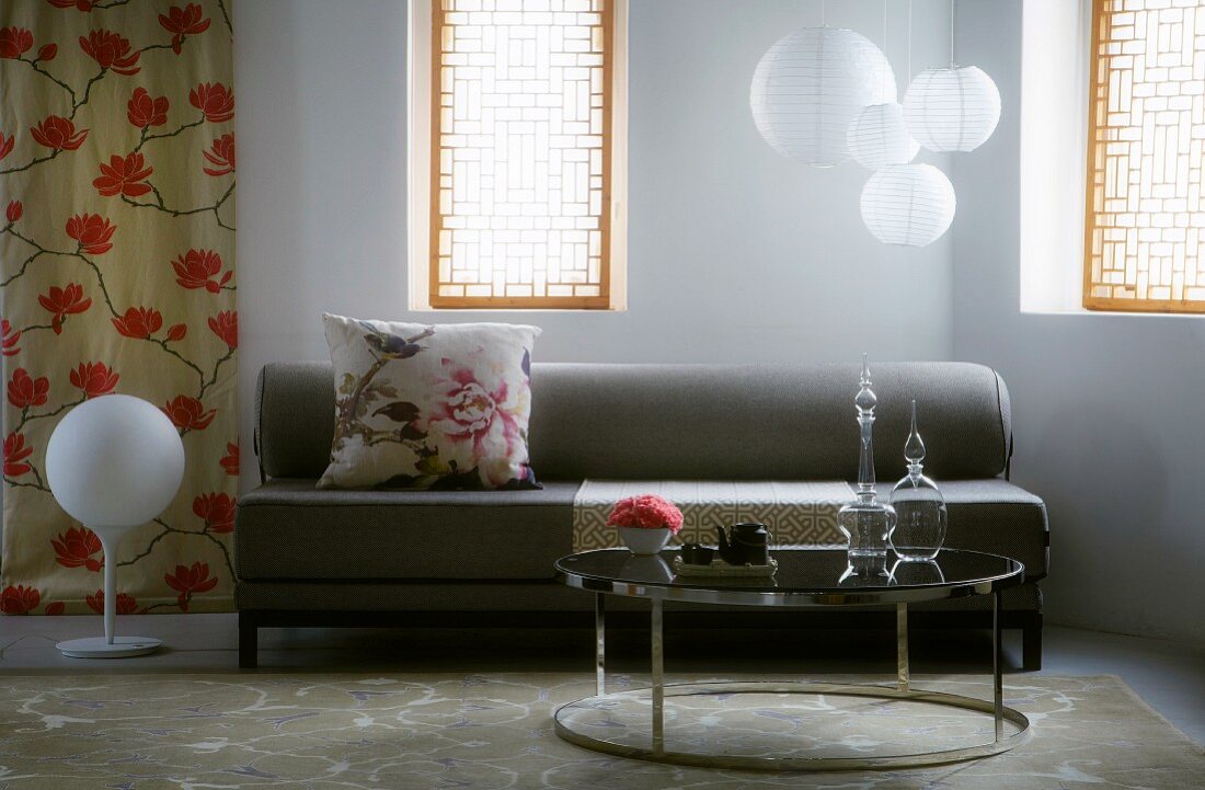 Retro-style, round side table in front of grey sofa and spherical pendant lamps between windows with Oriental-style screens