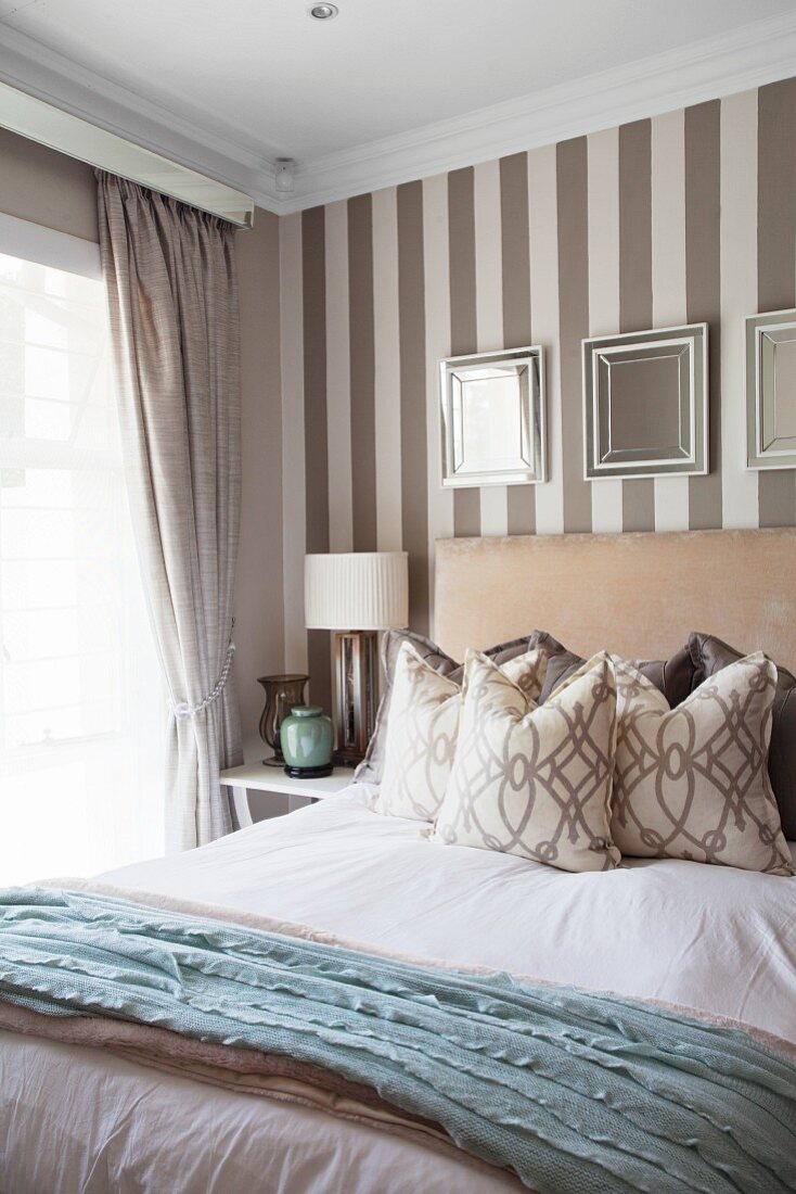 Framed mirrors on elegant wallpaper with broad stripes behind double bed with upholstered headboard