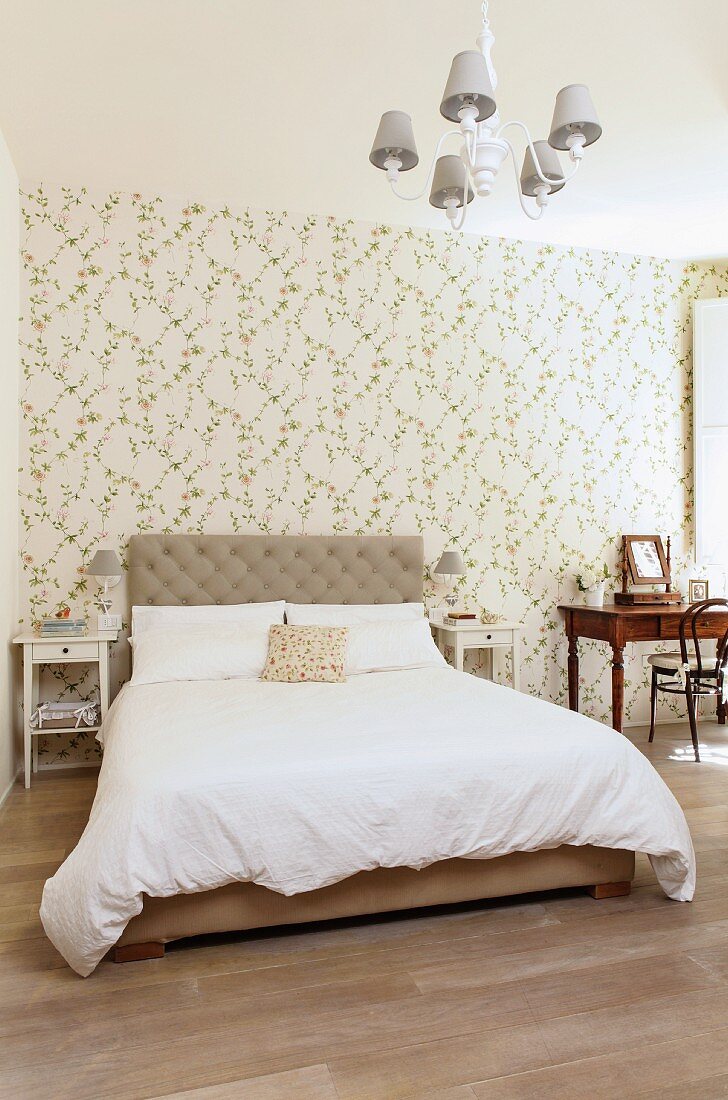 Double bed with upholstered headboard against floral wallpaper and antique dressing table a small distance away