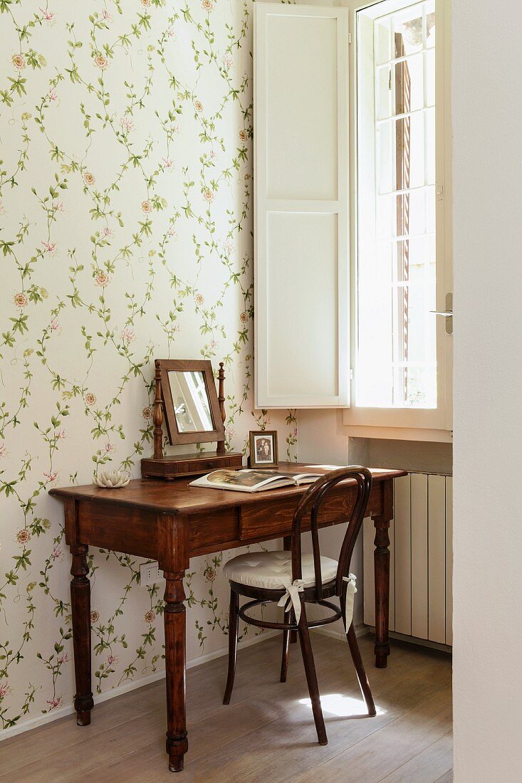 Antique writing desk with mirror and Thonet chair against wall with floral wallpaper below window with white interior shutters