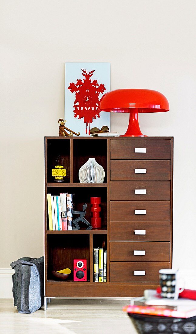 Bright red, 70s designer lamp and stylised cuckoo clock on cabinet with ornaments on shelves and drawers