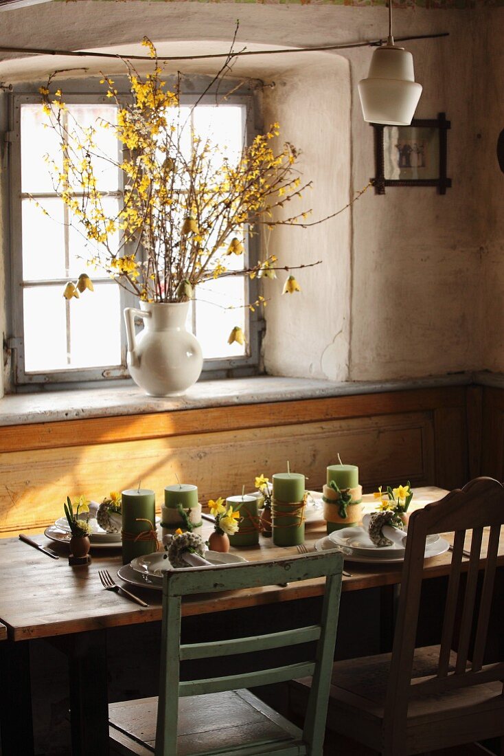 Frühlingshafter Esstisch mit Kerzen und Blumen vor Fenster, Forsythien in Porzellankrug auf Fensterbank