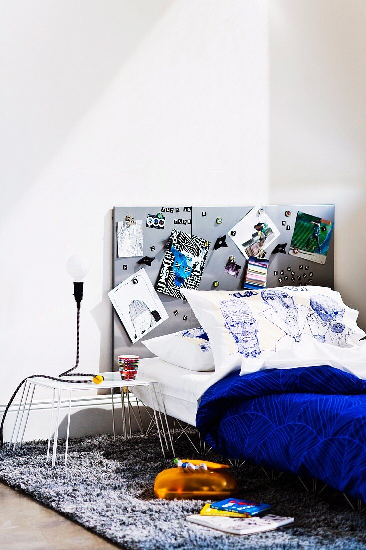 Teenager's bed with grey magnetic pinboard on wall, pillow painted with faces and blue bedspread next to table lamp on small metal table