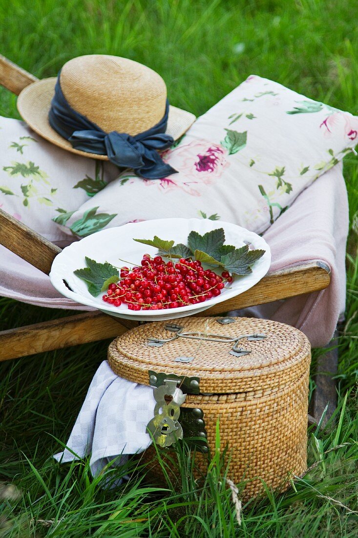 A deckchair with cushions, a straw hat and redcurrants, next to a picnic basket