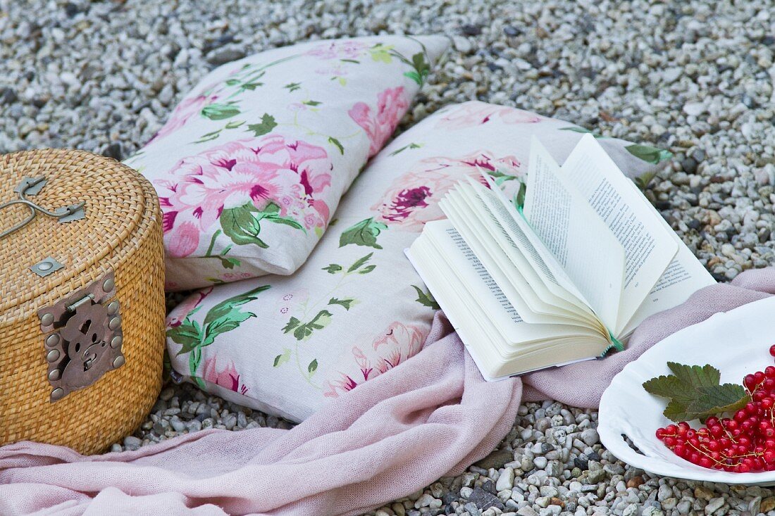 Kissen mit Blumenmuster, Decke, Buch und Picknickkorb auf Kiesboden