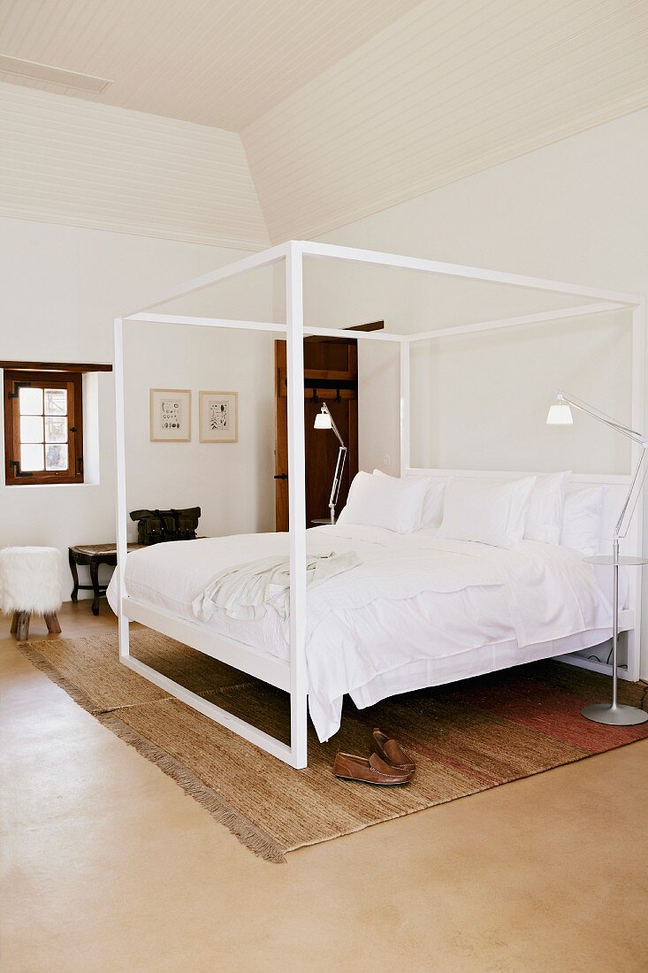 Canopied bed with white metal frame and white bed linen in simple bedroom with pale brown rug on lino-style floor