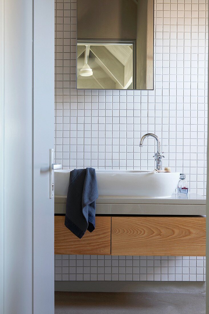 View through open bathroom door of washstand with oval sink and wooden-fronted drawers; mirrored cabinet on back wall with white mosaic tiles