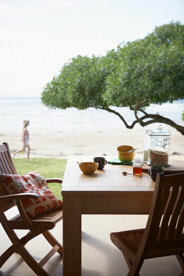 Zwei Holzstühle und Tisch mit Frühstücksutensilien auf Terrasse mit Blick auf Baumkrone und Meeresstrand