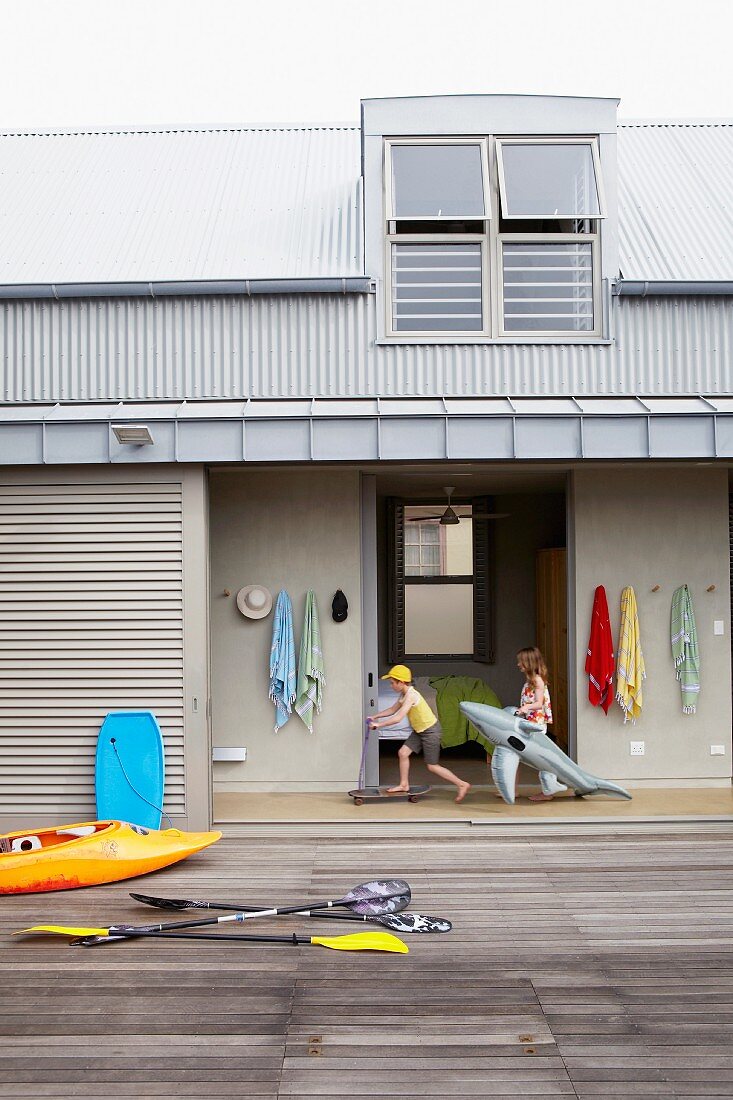 Holzterrasse mit Paddel und Boot vor Strandhaus mit Blechdach im Industriestil; spielende Kinder und Badeutensilien im offenen Flurbereich