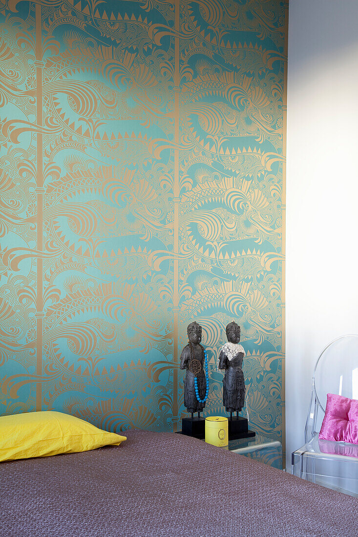 Bed with purple bed linen, patterned wallpaper and Asian figures on bedside table