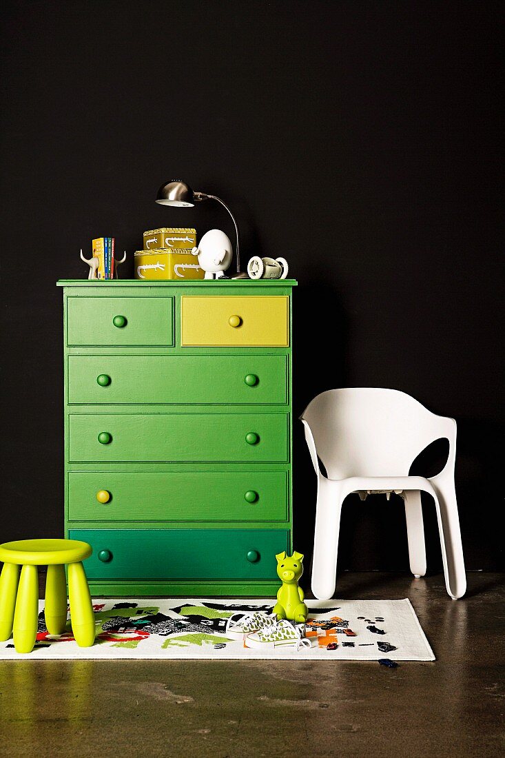 Grüne Schubladenkommode an schwarzer Wand; daneben ein weisser Kunststoffstuhl und ein Kinderhocker
