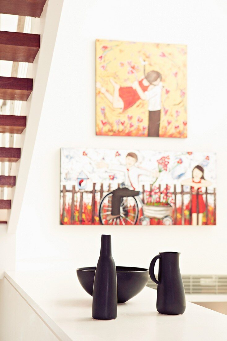 Stillleben mit schwarzen Tongefässen auf halbhohem Schrank vor Treppennische und Bilder mit Kindermotiven an Wand