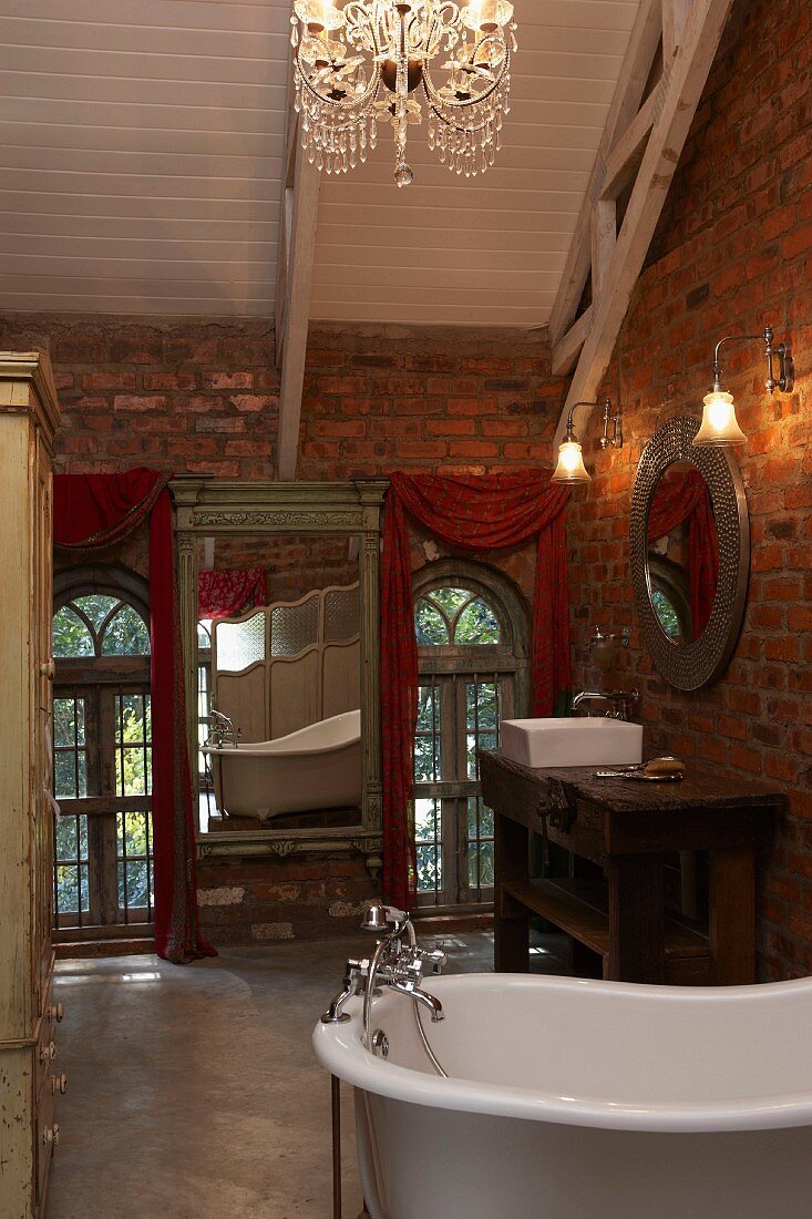 Antique mirror on wall and free-standing bathtub in romantic, vintage bathroom in attic interior with brick walls