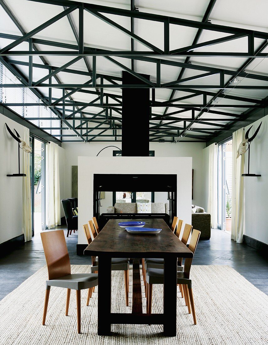 Rustikale Tafel aus dunklem Holz und Stühle im Retro-Stil auf Teppich in hallenartigem Wohnraum mit sichtbarer Dachkonstruktion; Raumteiler vor Loungebereich