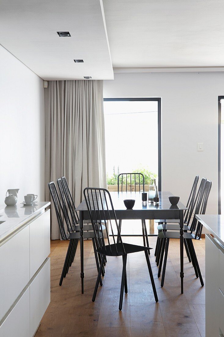 View from white fitted kitchen with wooden floor into open-plan dining area with dark, designer chairs