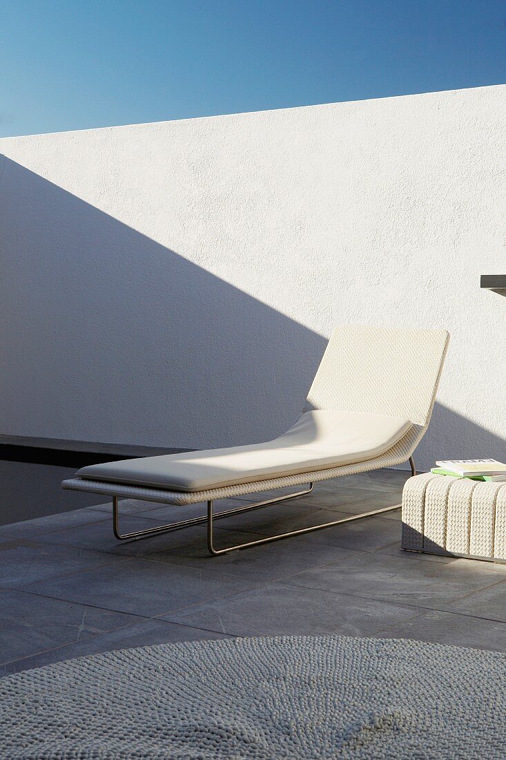 Elegant, white designer lounger on grey, stone flags of terrace against white wall of sunny courtyard