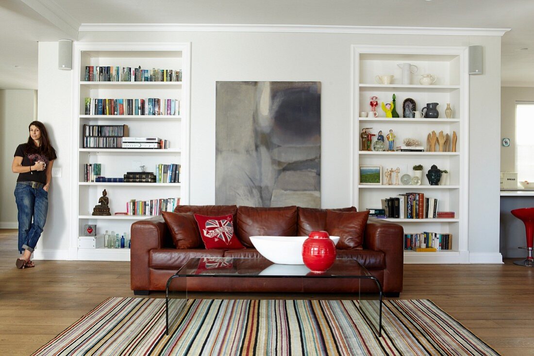 Plexiglass coffee table on striped rug and leather couch in front of fitted shelving; woman in background