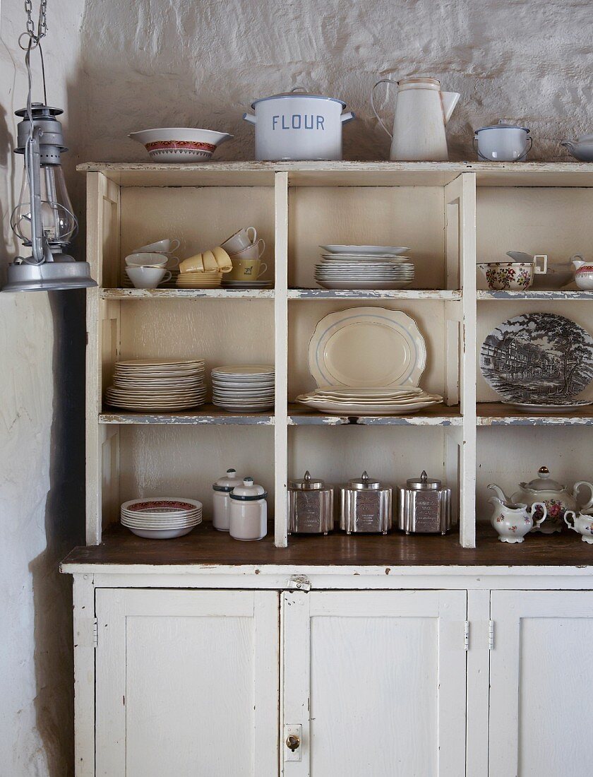Simple, white kitchen dresser with crockery in open-fronted shelves against rustic, plastered wall