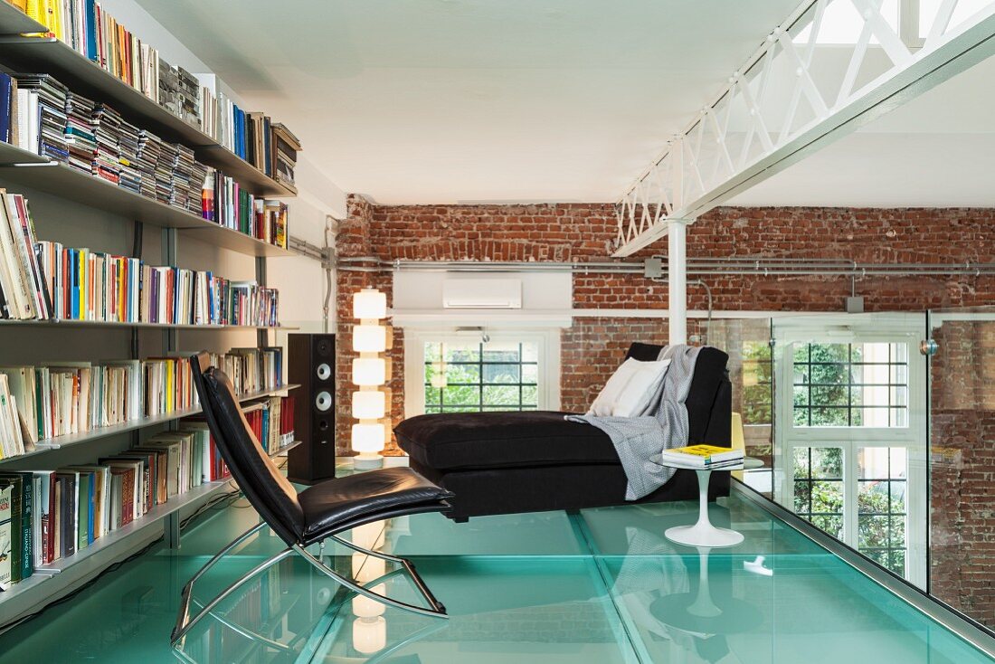 Loft apartment with steel girders and brick walls; leather armchair in front of bookcase on gallery bedroom with glass floor