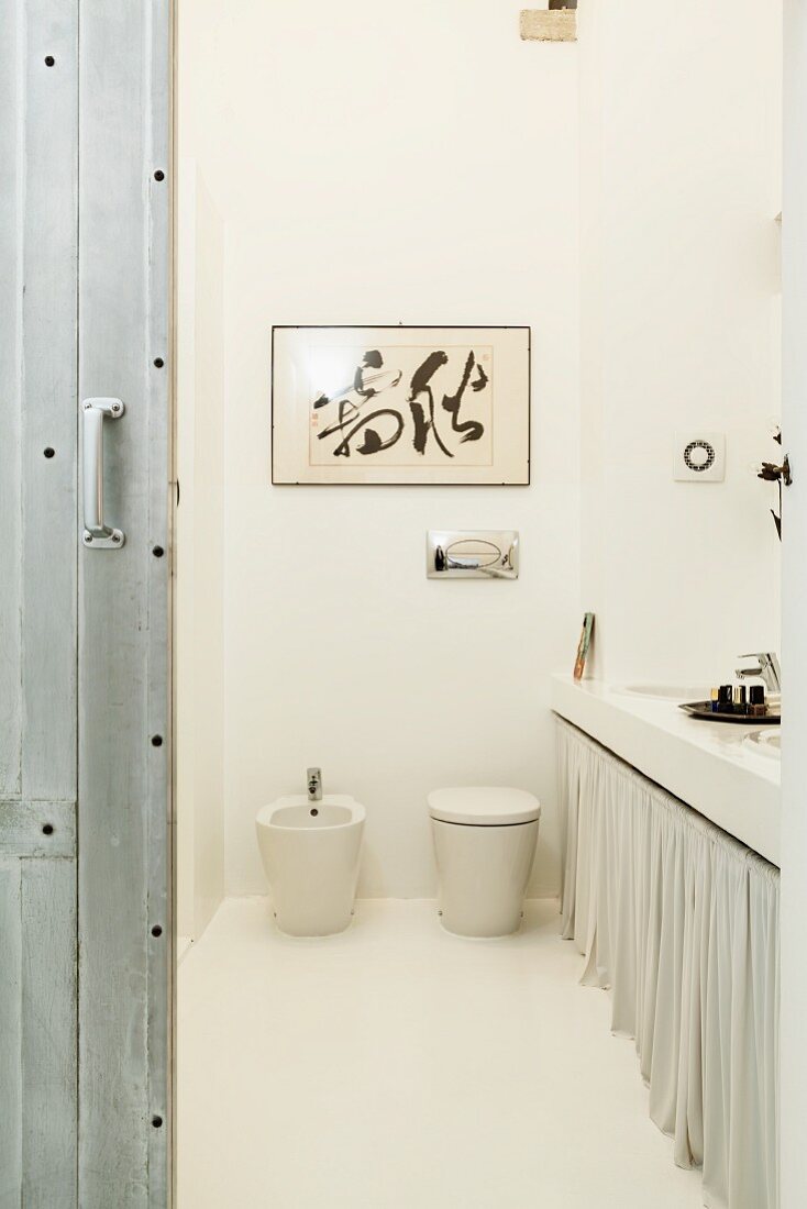 Helle Badgestaltung im Loft mit abstrakter Malerei über WC und Bidet, daneben Waschtischzeile mit Vorhang und Stahlschiebetür im Vordergrund