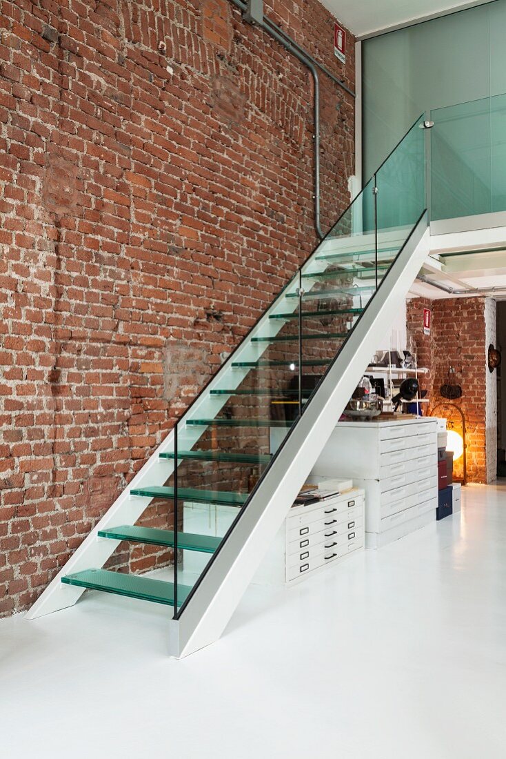 Treppenaufgang mit Glasbrüstung und Glasstufen vor unverputzter Backsteinwand in Designer-Loft mit weißem Fussboden; Planschränke unter Treppenlauf