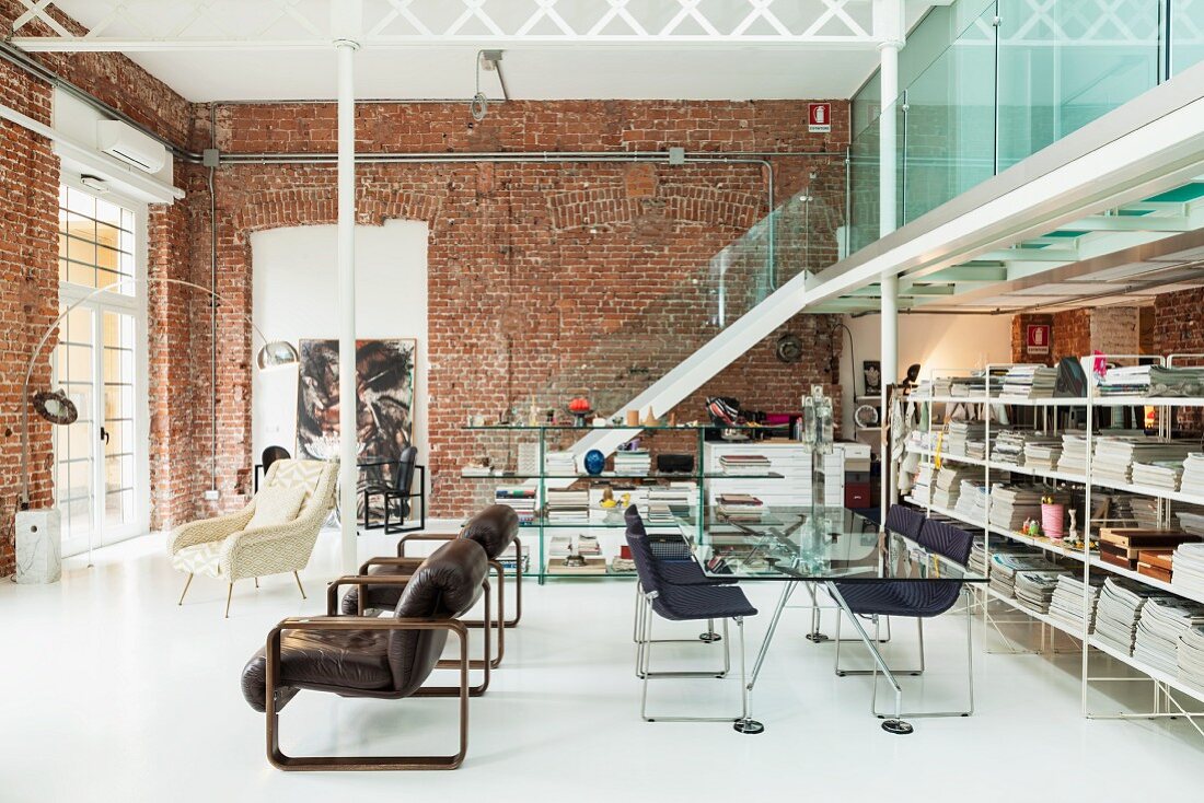 Designer-Loft mit Stahllederstühlen an Glastisch zwischen Bücherwand und Retrosesseln; im Hintergrund eine Galerietreppe mit kühler Glasbrüstung vor alter Backsteinwand