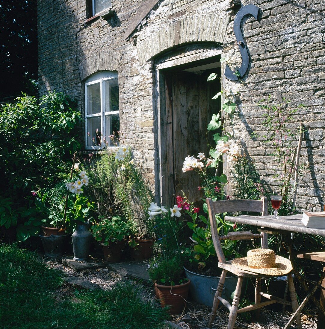 Außenansicht eines rustikalen Landhauses aus Stein mit einem Holztisch, Stuhl, blühenden Blumen und offener Tür