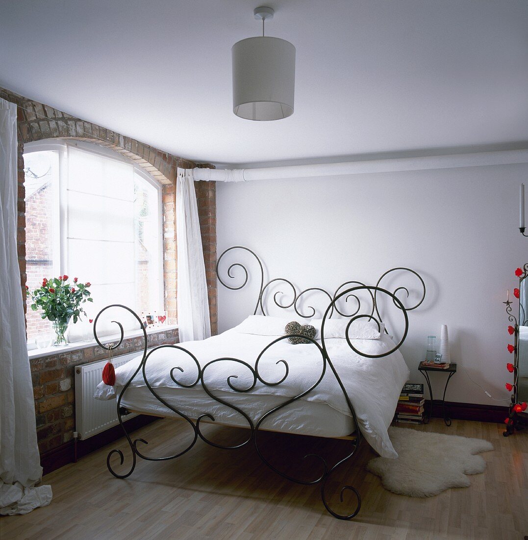 Schlafzimmer mit dekorativem schmiedeeisernen Bett, Backsteinwand, Holzfußboden und gewölbtem, sonnigen Fenster