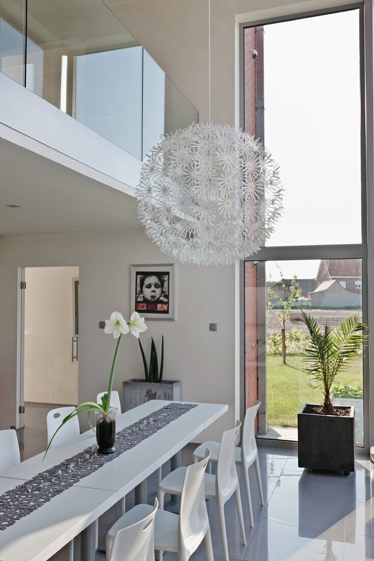 Spherical pendant lamp with white flower details above modern, white dining area with glossy floor below gallery with glass balustrade