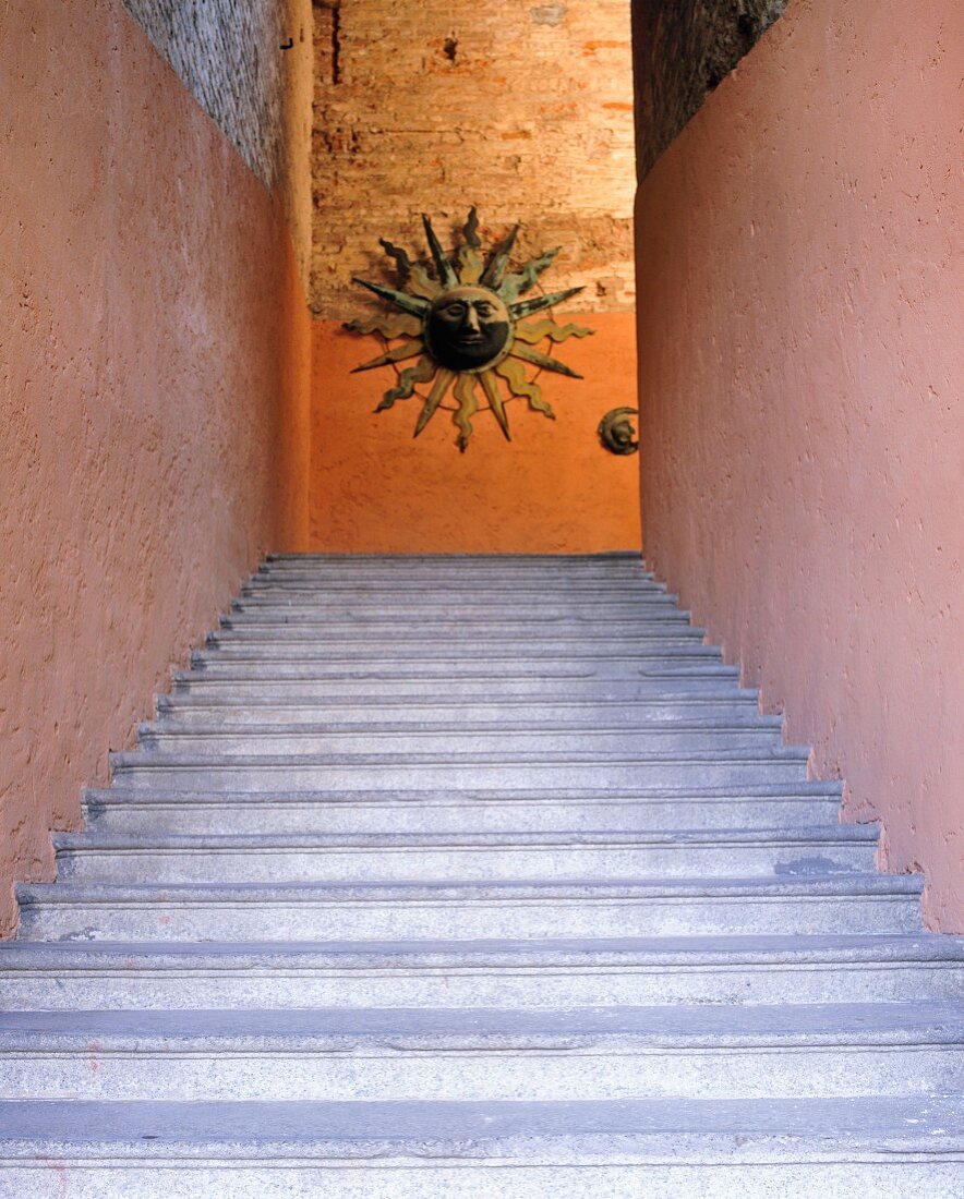 Blick von unten auf Steintreppenaufgang und orange getönte Wand mit Sonnenskulptur