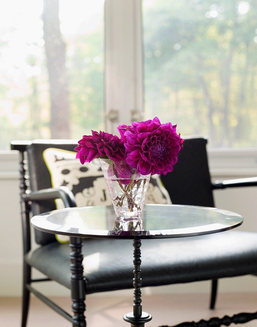 Violette Dahlien in Glasvase auf Beistelltisch und Stuhl vor dem Fenster