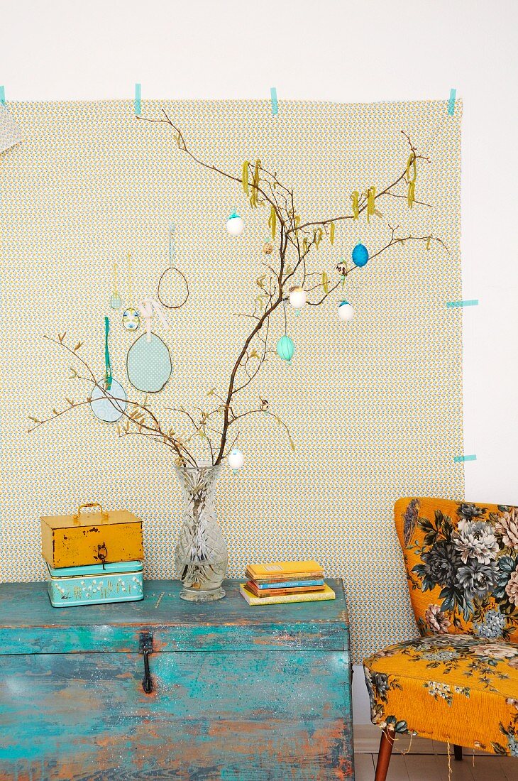 Branches hung with painted Easter eggs in glass vase on vintage wooden trunk next to armchair with 50s-style patterned upholstery