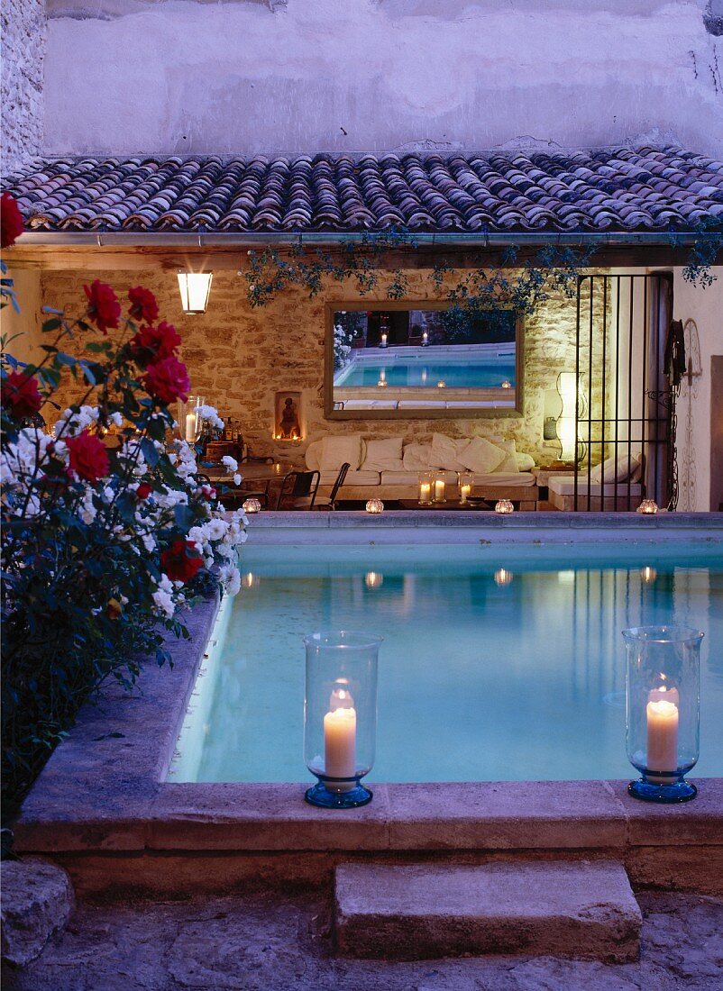 Romantische Dämmerstimmung mit Windlichtern an Pool-Umrandung; Sitzplatz vor rustikalem Landhaus im Hintergrund