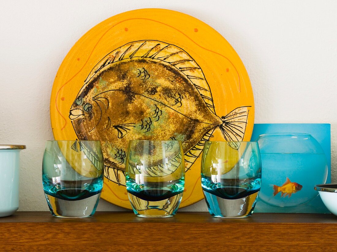 Drei blau schimmernde Trinkgläser vor gelber Platte mit Fischmotiv und daneben eine Abbildung mit Fisch im Wasserglas