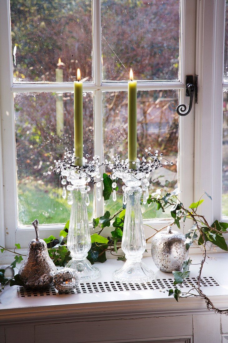 Brennende Kerzen in Glasleuchtern, versilberter Apfel und Birne zwischen Efeuranken auf einem Fensterbrett