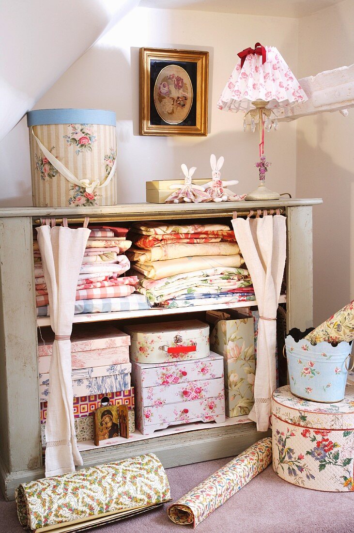 Florale Muster im romantischen Landhausstil - Schachteln, Lampenschirm und gefaltete Wäsche in Regal mit drapiertem Vorhang