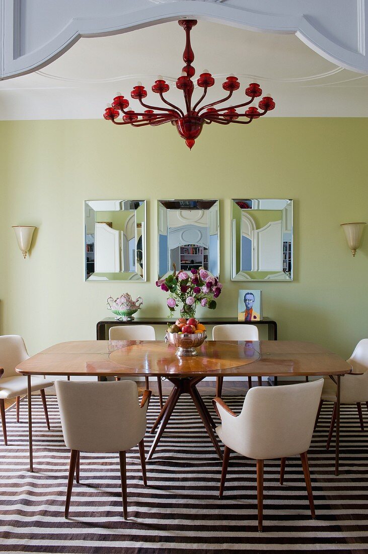 Large, square, exotic wood dining table, retro chairs with pale upholstery, red chandelier and framed mirrors on painted wall in spacious dining room