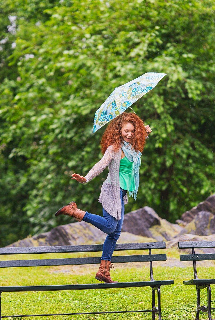 Frau hüpft mit Regenschirm auf Parkbank herum