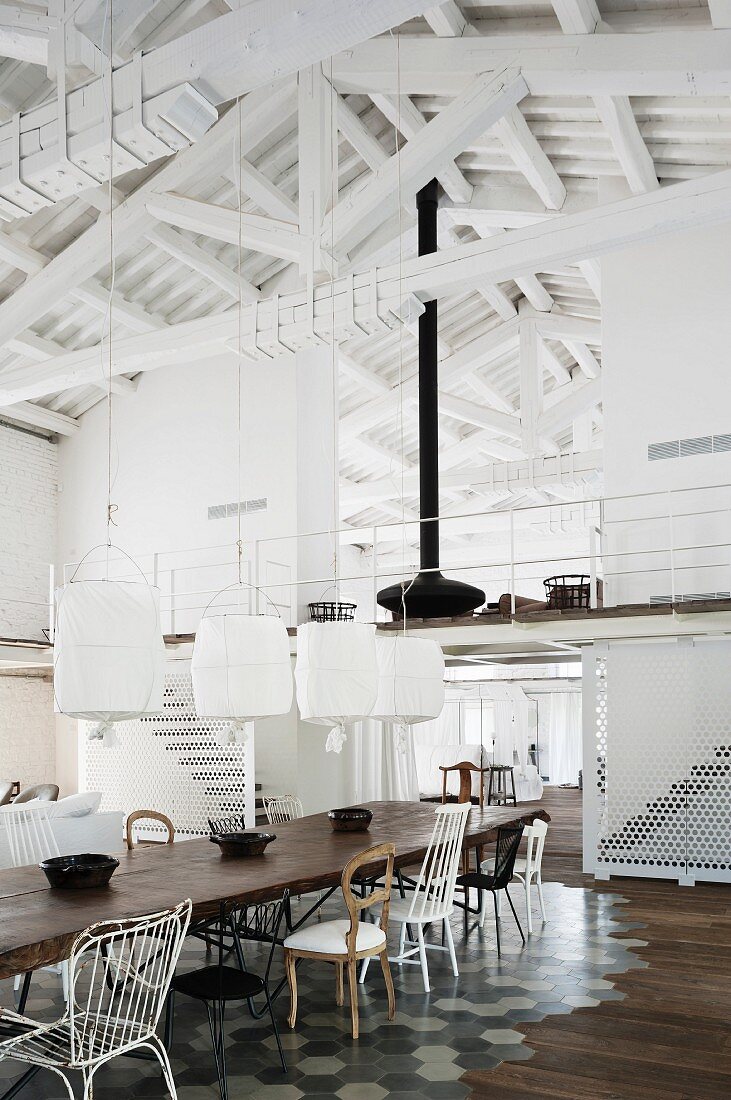 Esstisch mit verschiedenen Stühlen auf gefliestem Bodenbereich und Hängeleuchten mit weissen Stoffschirmen vor Galerie im Dachgeschoss mit geweisseltem Dachstuhl