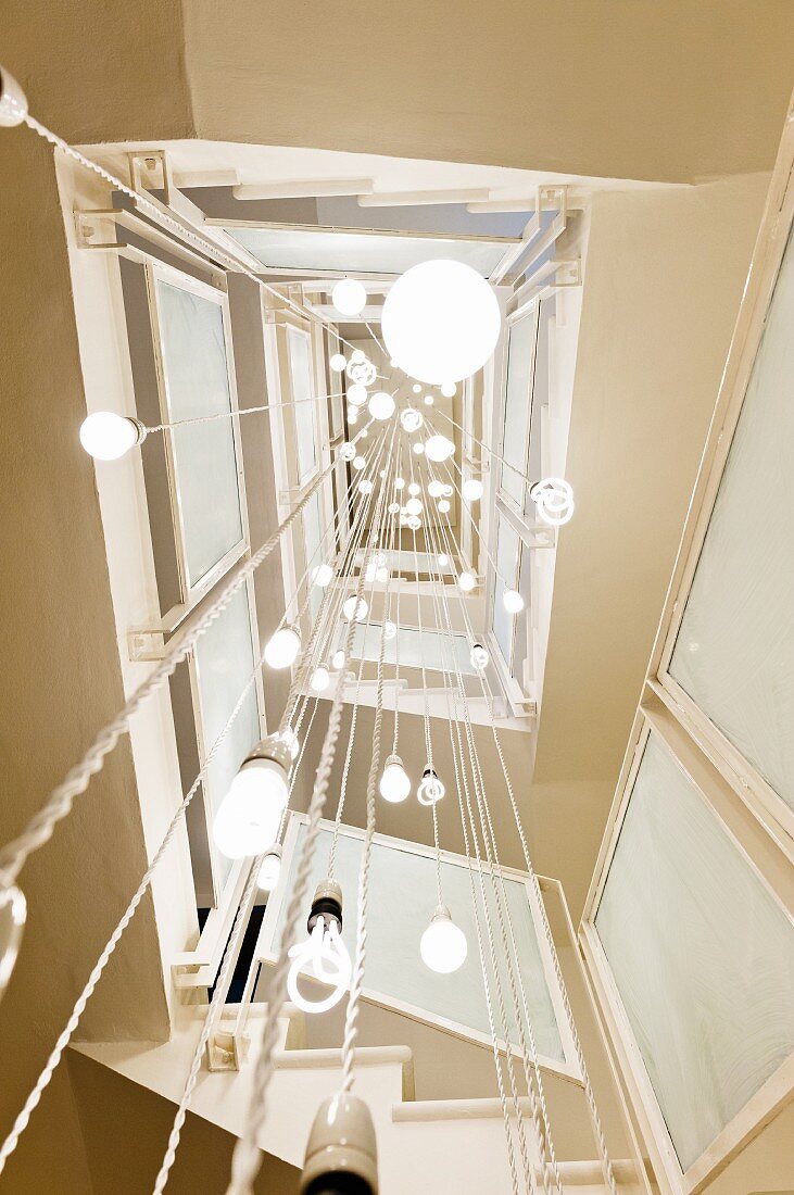 Blick von unten ins Treppenauge mit von Decke abgehängten, verschiedenen Hängeleuchten als Lichtskulptur