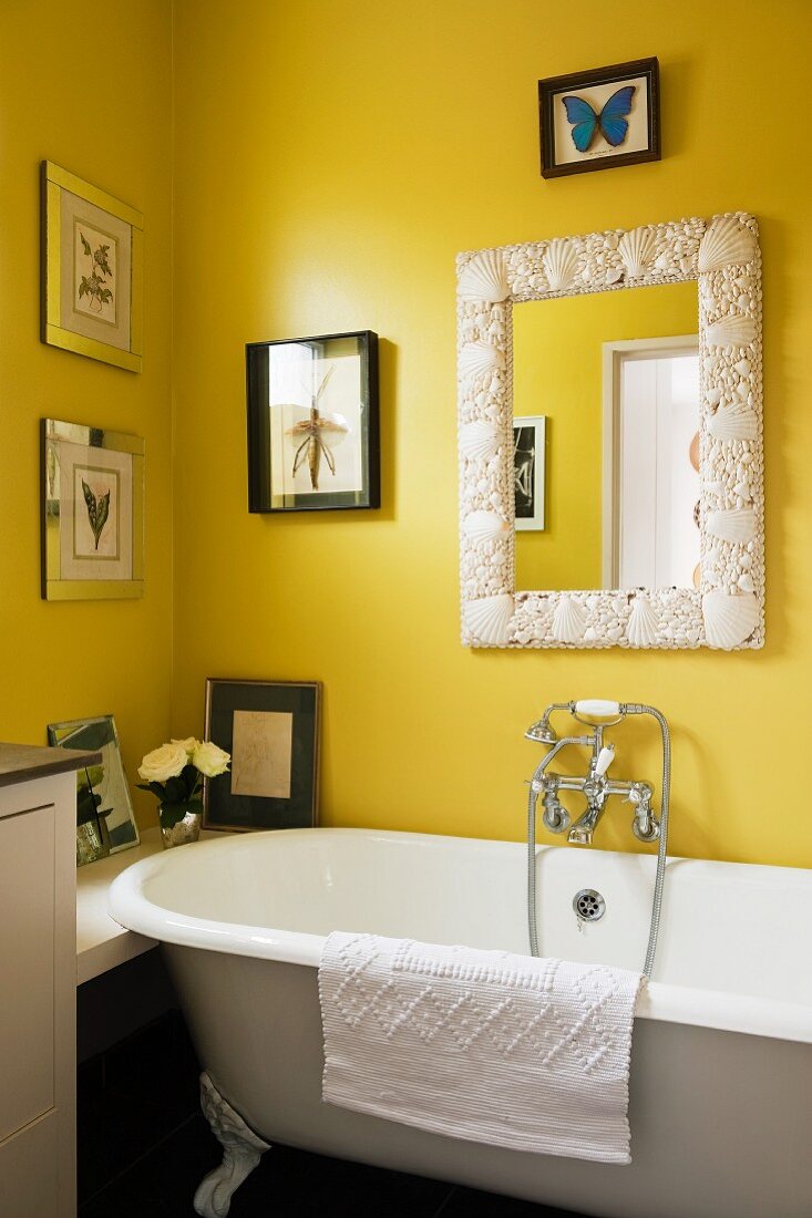 Nostalgische Badewanne an gelber Wand mit Muscheln verziertem Spiegelrahmen und Insektenbildern
