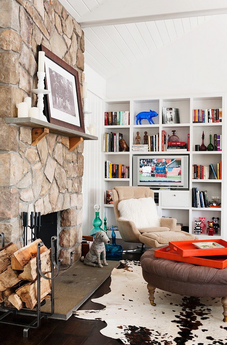 Seating area with armchair, ottoman, cowhide rug and white fitted shelves in front of rustic, stone chimney breast