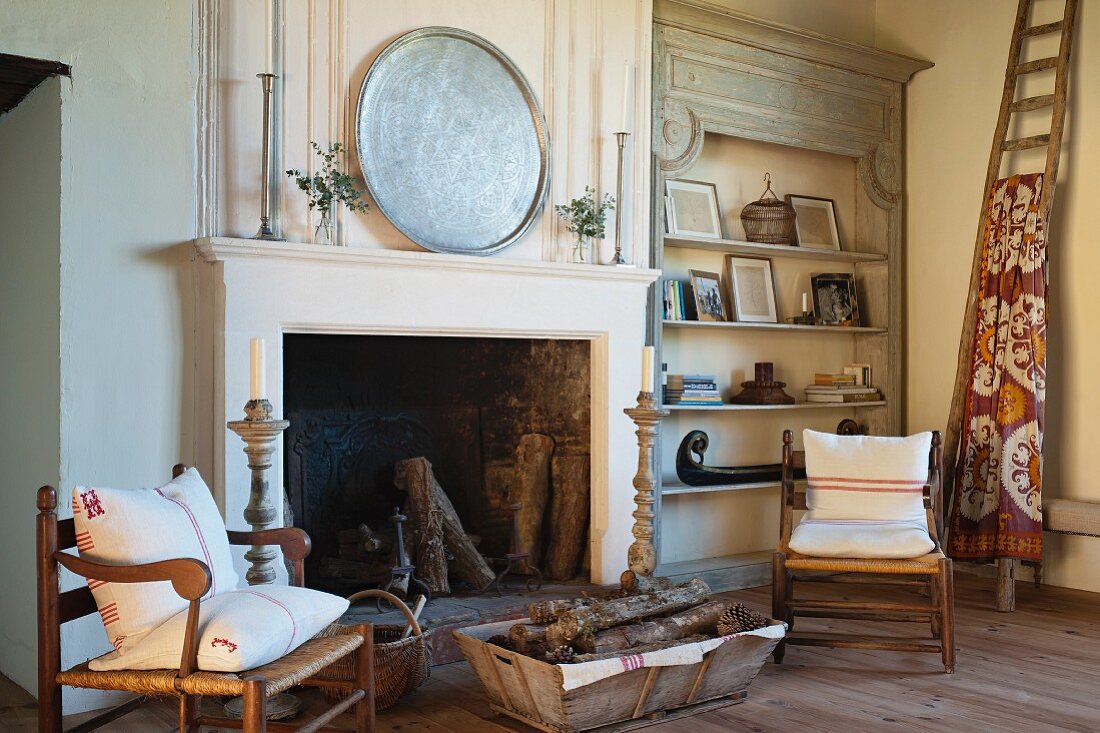 Rush-bottom armchairs with wooden frames flanking open fireplace and antique wooden shelves in rustic living room