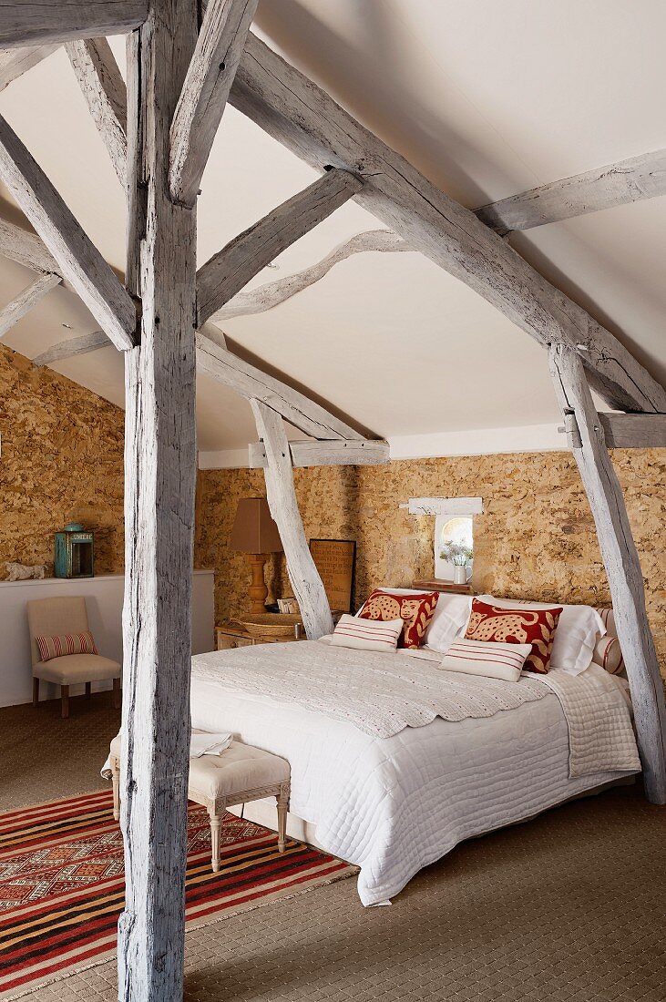 Rustic attic bedroom with exposed wooden roof structure, stone walls and Moroccan rug next to bed