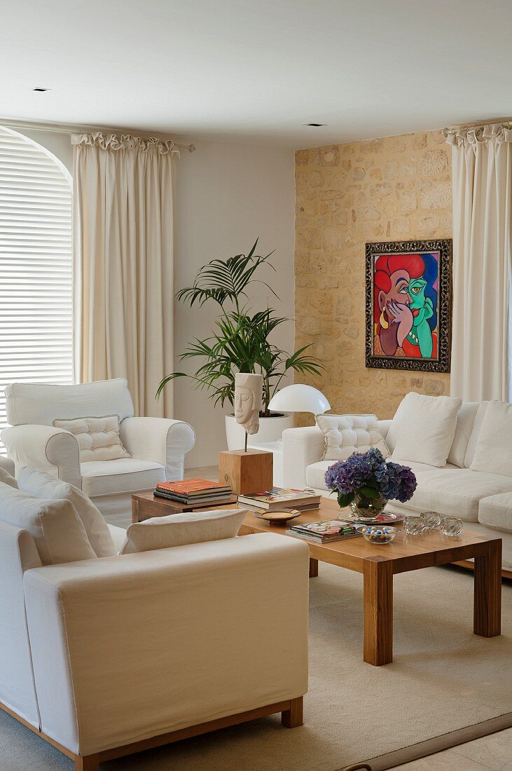 Sitzgruppe mit hellen Polstersofas und Holztisch im Wohnzimmer; bunte Malerei an Natursteinwand im Hintergrund