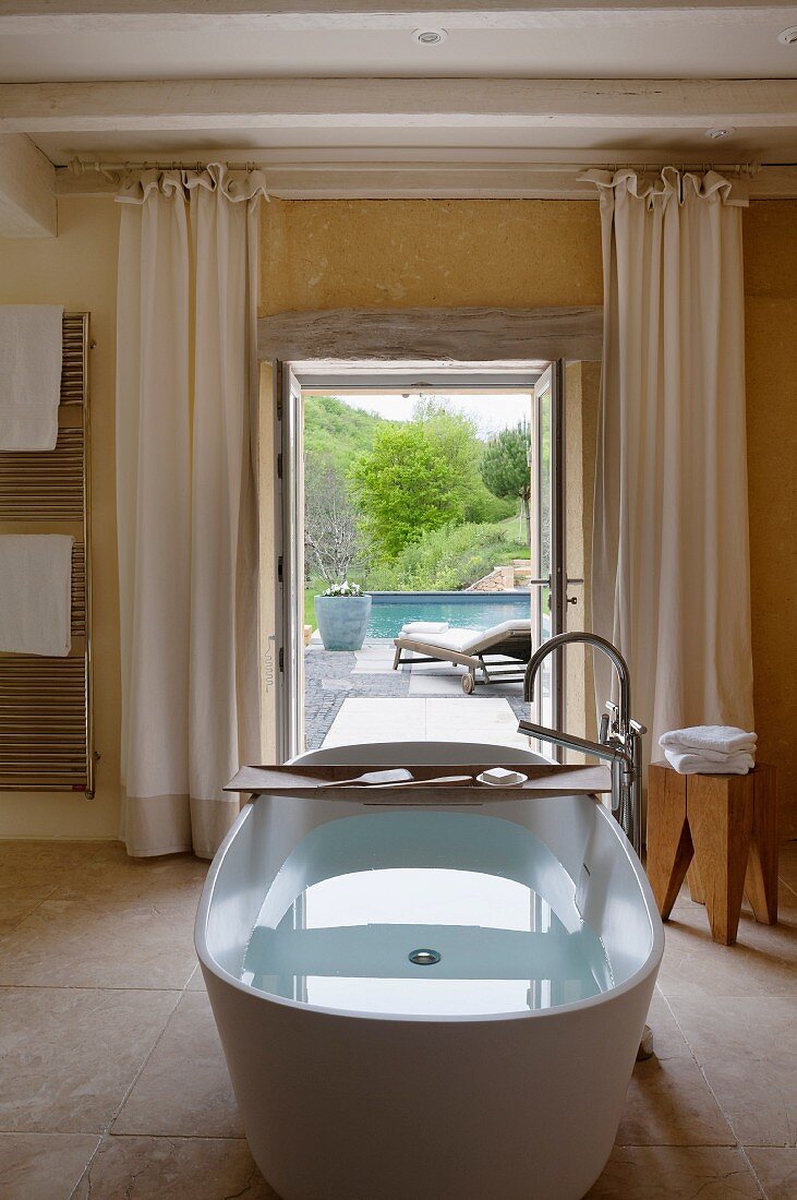 Free-standing bathtub with wooden shelf; open terrace door with view of pool in background